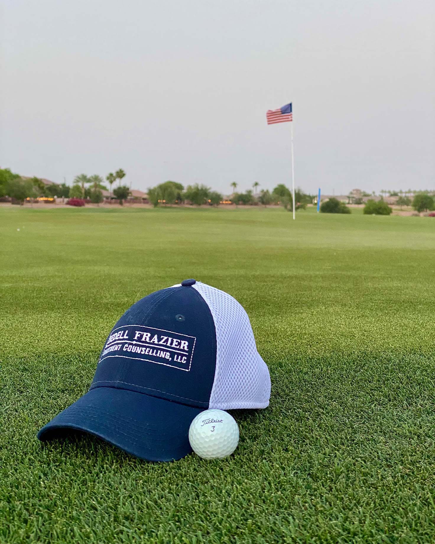 Bedell Frazier branded navy blue and white trucker hat placed on a golf course green alongside a Titleist golf ball, with a flagpole displaying the American flag in the background.