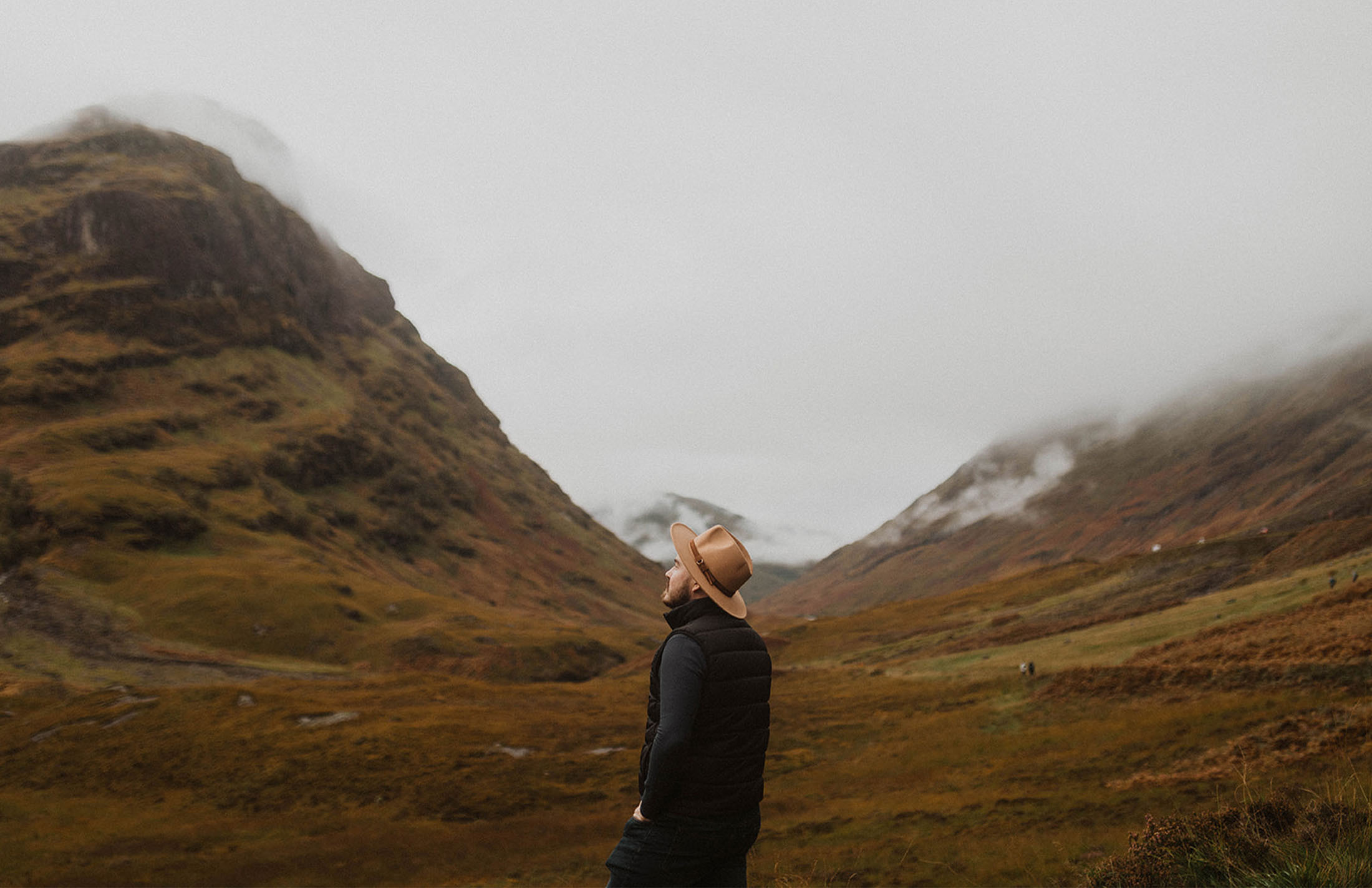 A person wearing a hat stands amidst the scenic Scottish Highlands under a cloudy sky, surrounded by green and brown rolling hills.