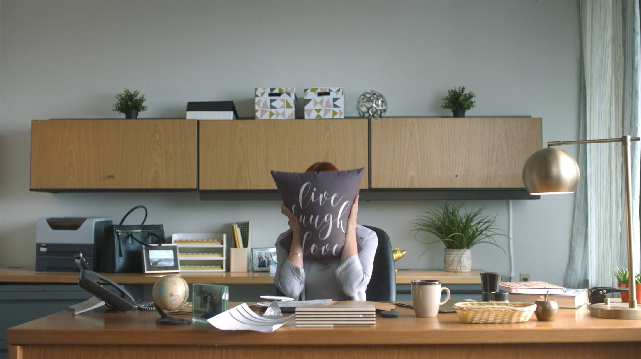 Person sitting at a neatly organized desk in a modern office, holding a decorative pillow with the phrase “Live Laugh Love,” showcasing a humorous and relatable workplace moment.