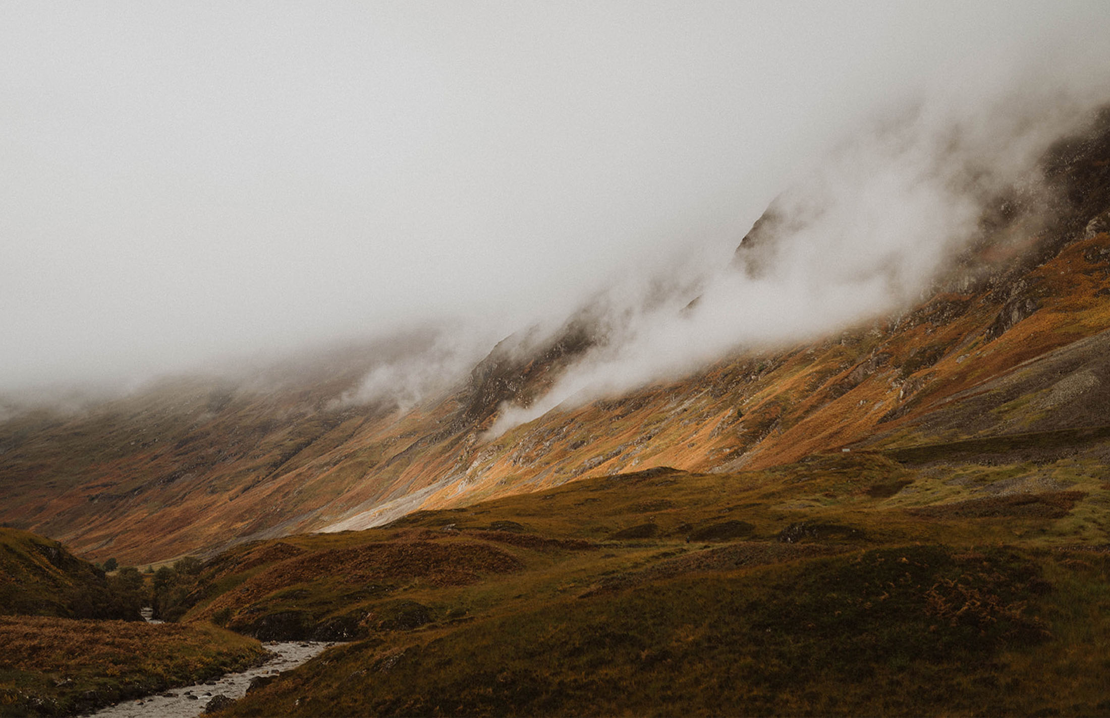 Cloud-covered Scottish Highlands featuring rolling green and orange hills with a stream running through the valley.