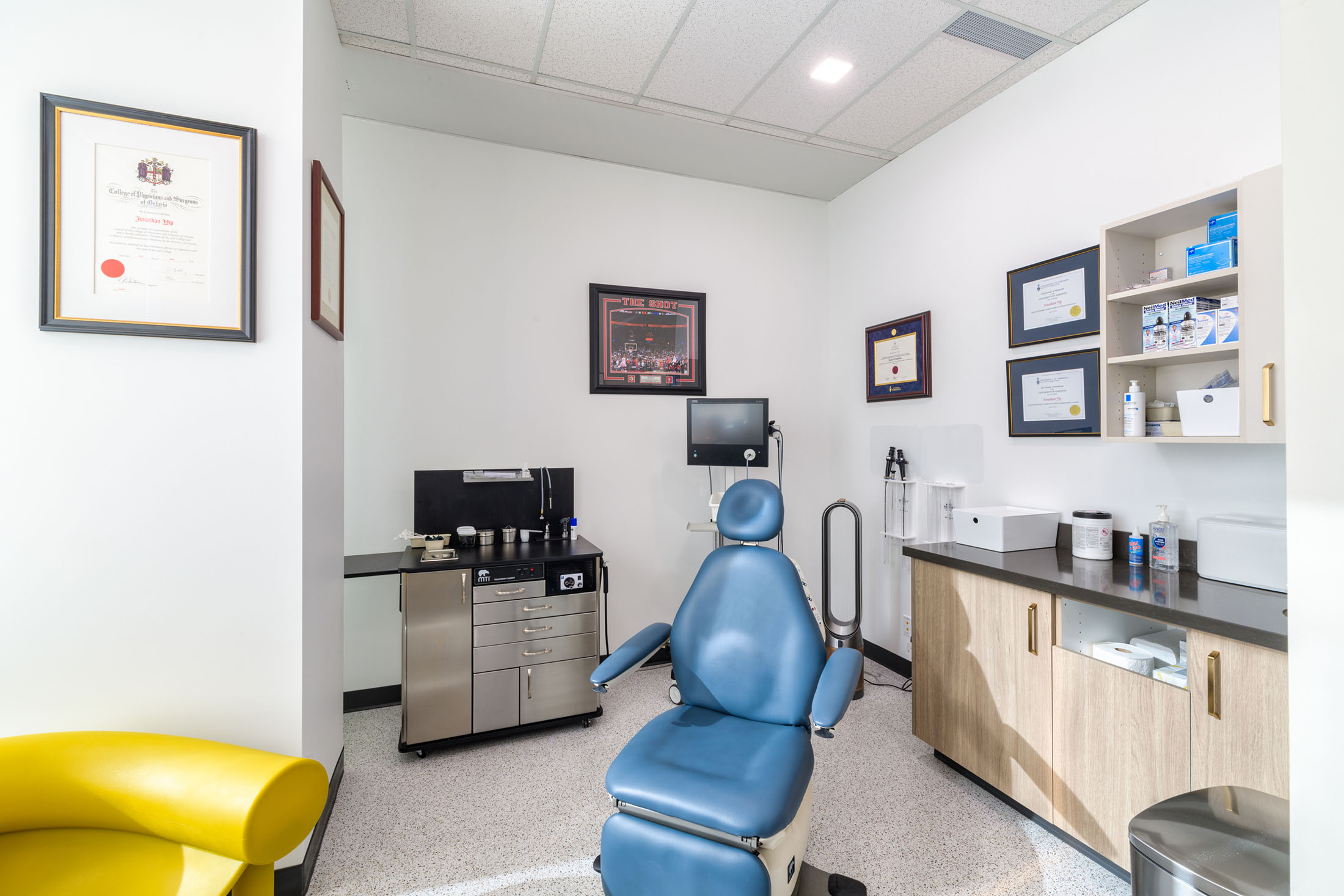 A clean and well-lit medical examination room featuring a blue patient chair, framed diplomas, and organized medical equipment on the counters and shelves.