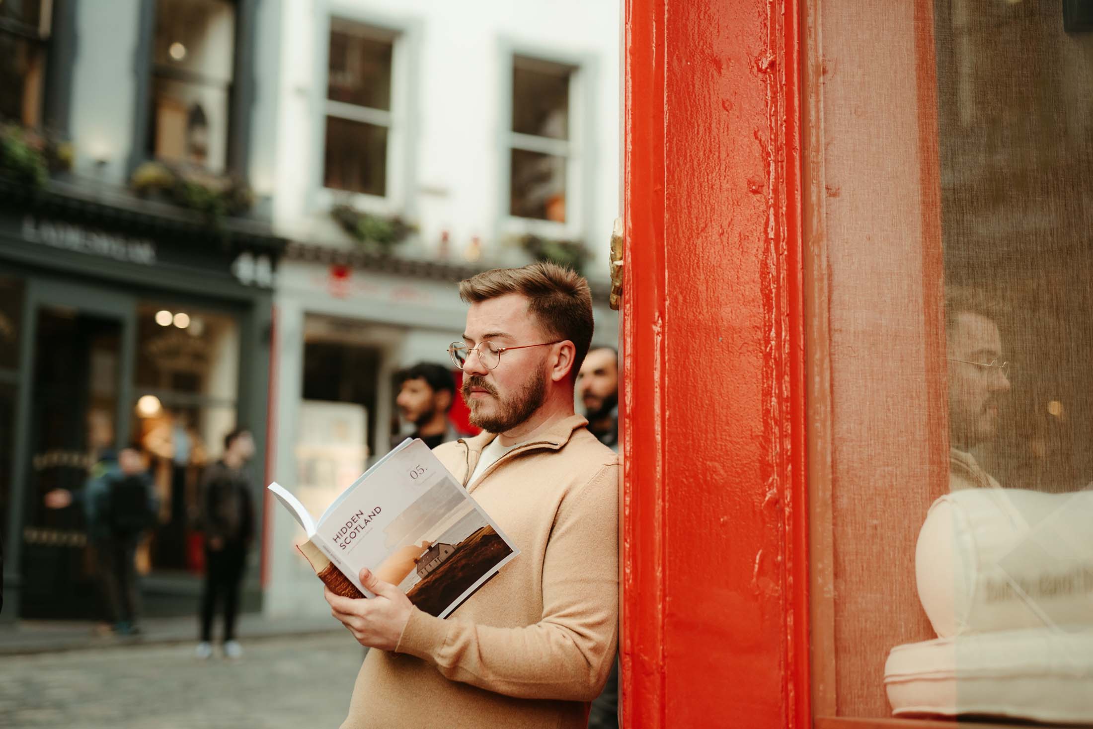 A man reading a Hidden Scotland travel guide in a city setting, leaning against a vibrant red door with a street view in the background.