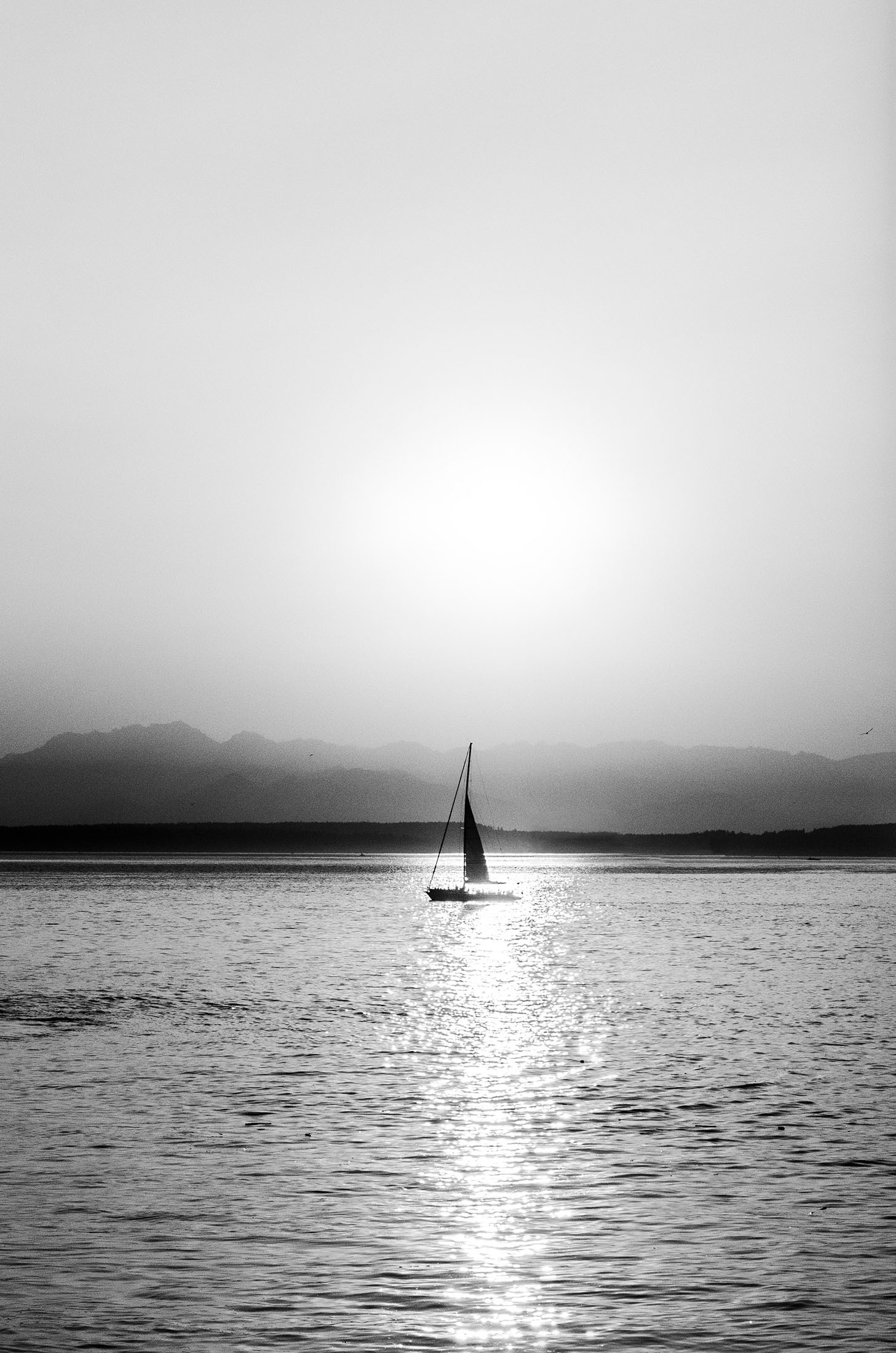 A black-and-white photo of a sailboat on a calm sea with mountains in the background, captured at sunset