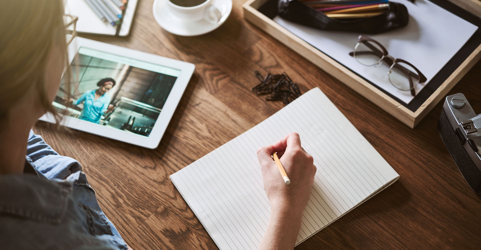 Overhead view of a creative professional writing in a notebook, surrounded by tools like a tablet, camera, and glasses, illustrating the process of crafting branding with words and visuals.