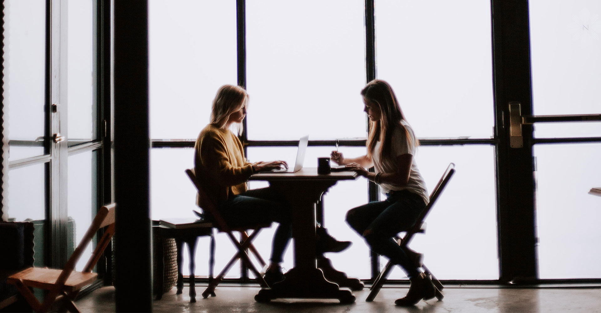 Two women collaborating at a table with laptops and notebooks, highlighting effective communication and teamwork in branding and website design projects.