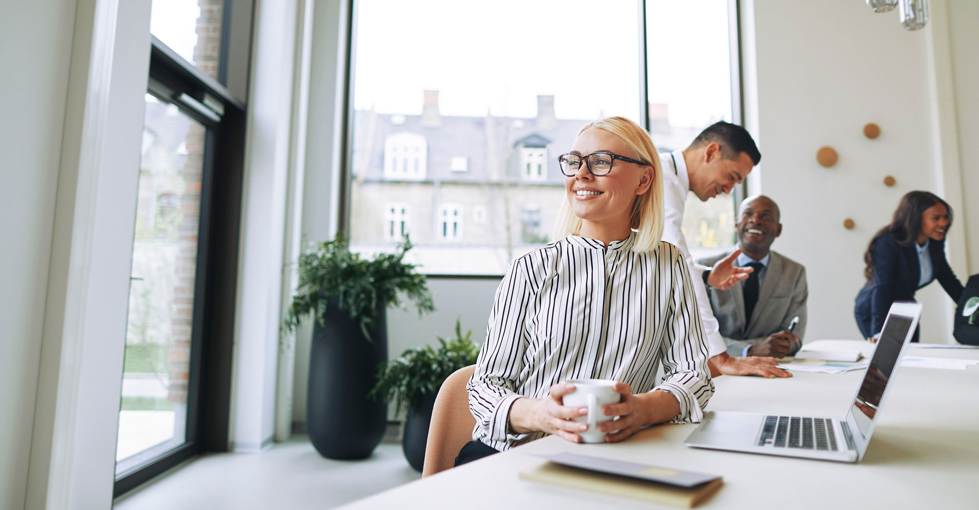 Confident professional woman holding a coffee cup at a modern workspace, surrounded by a collaborative team, emphasizing the value of investing in brand development and design.