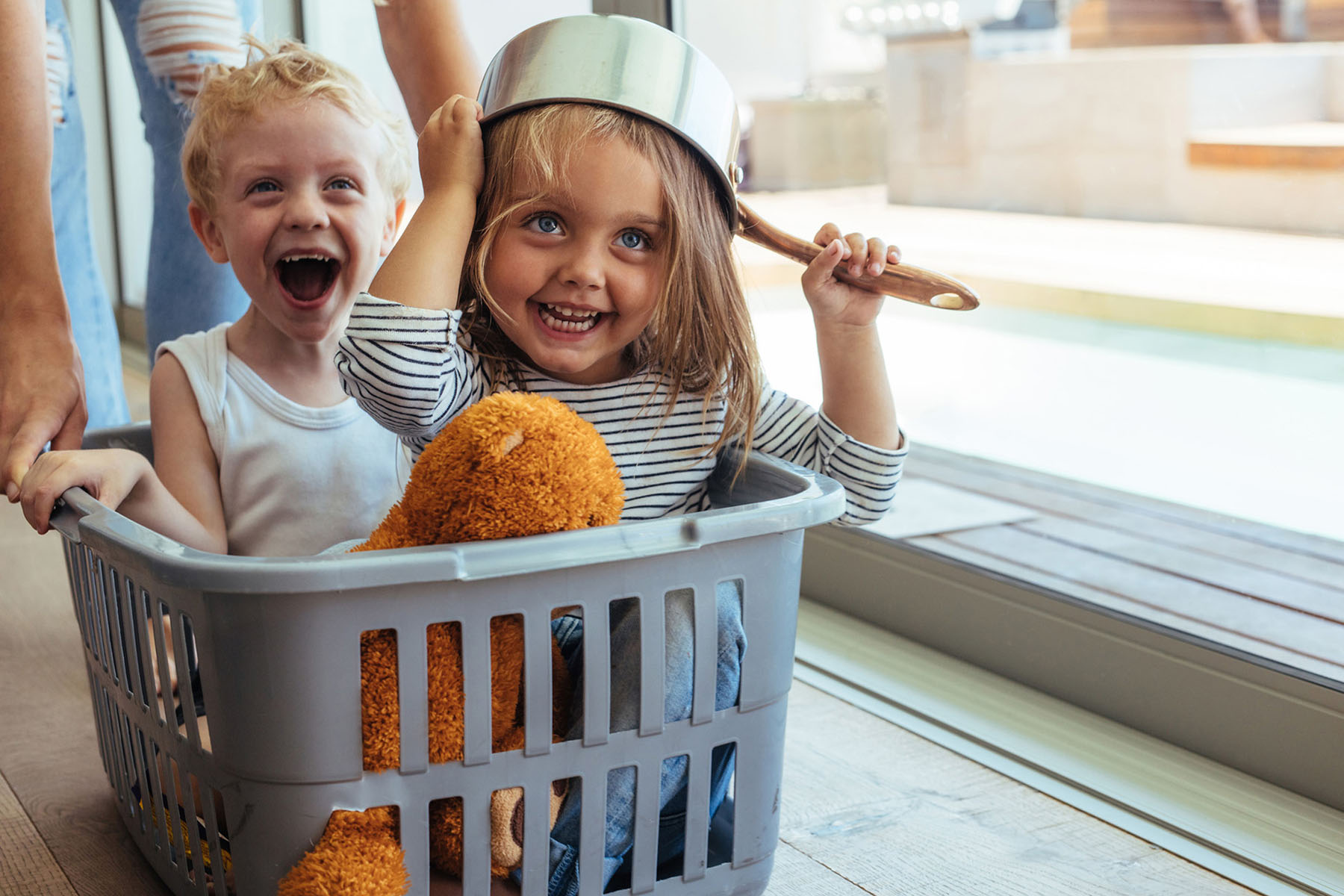 Children playing indoors, one wearing a pot on their head as a hat and the other laughing, capturing a joyful and playful moment, representing the trusted and caring childcare services of Nannies by Noa.