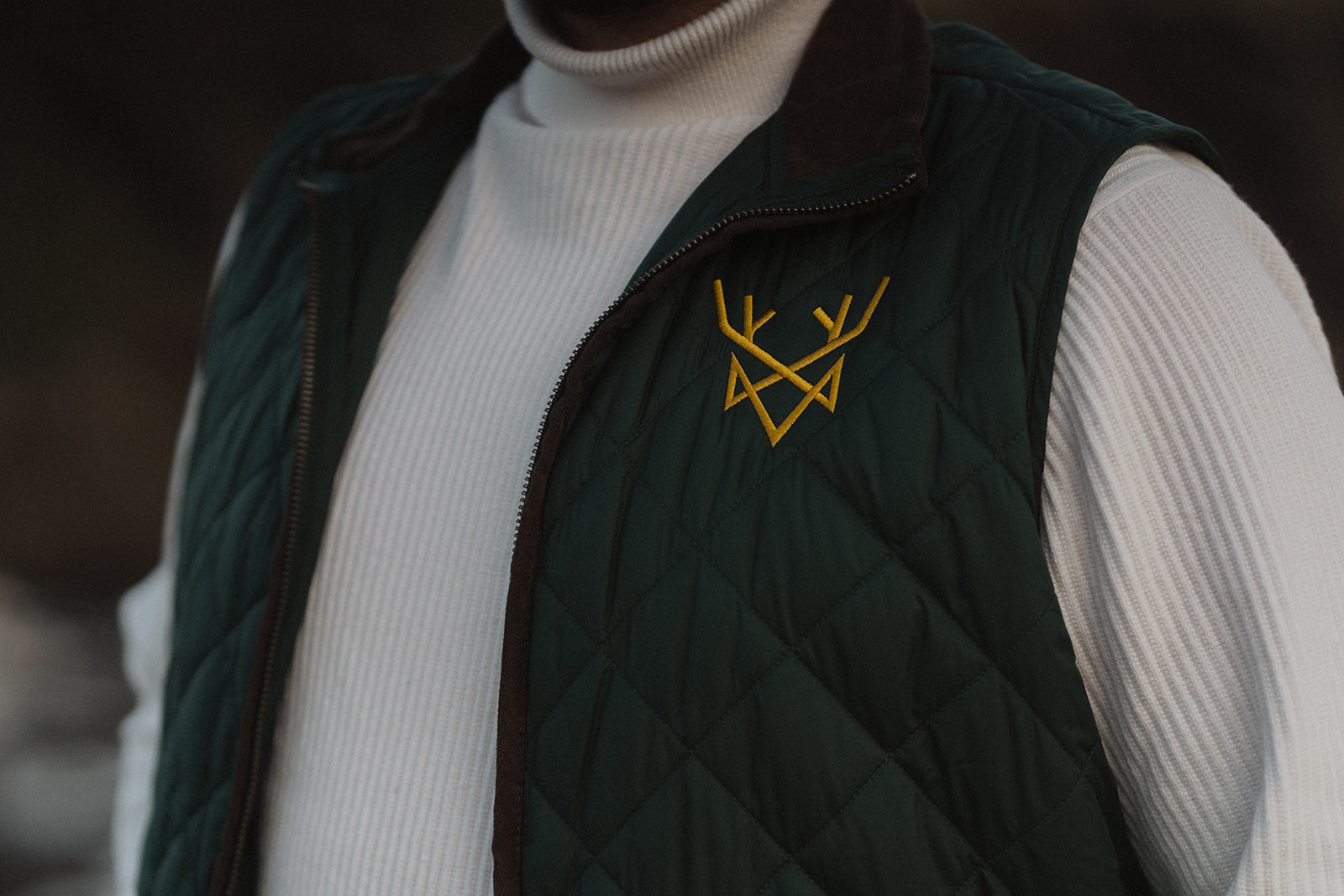 Royal B Travel branded vest in dark green with the logo embroidered in gold, worn over a white ribbed sweater.