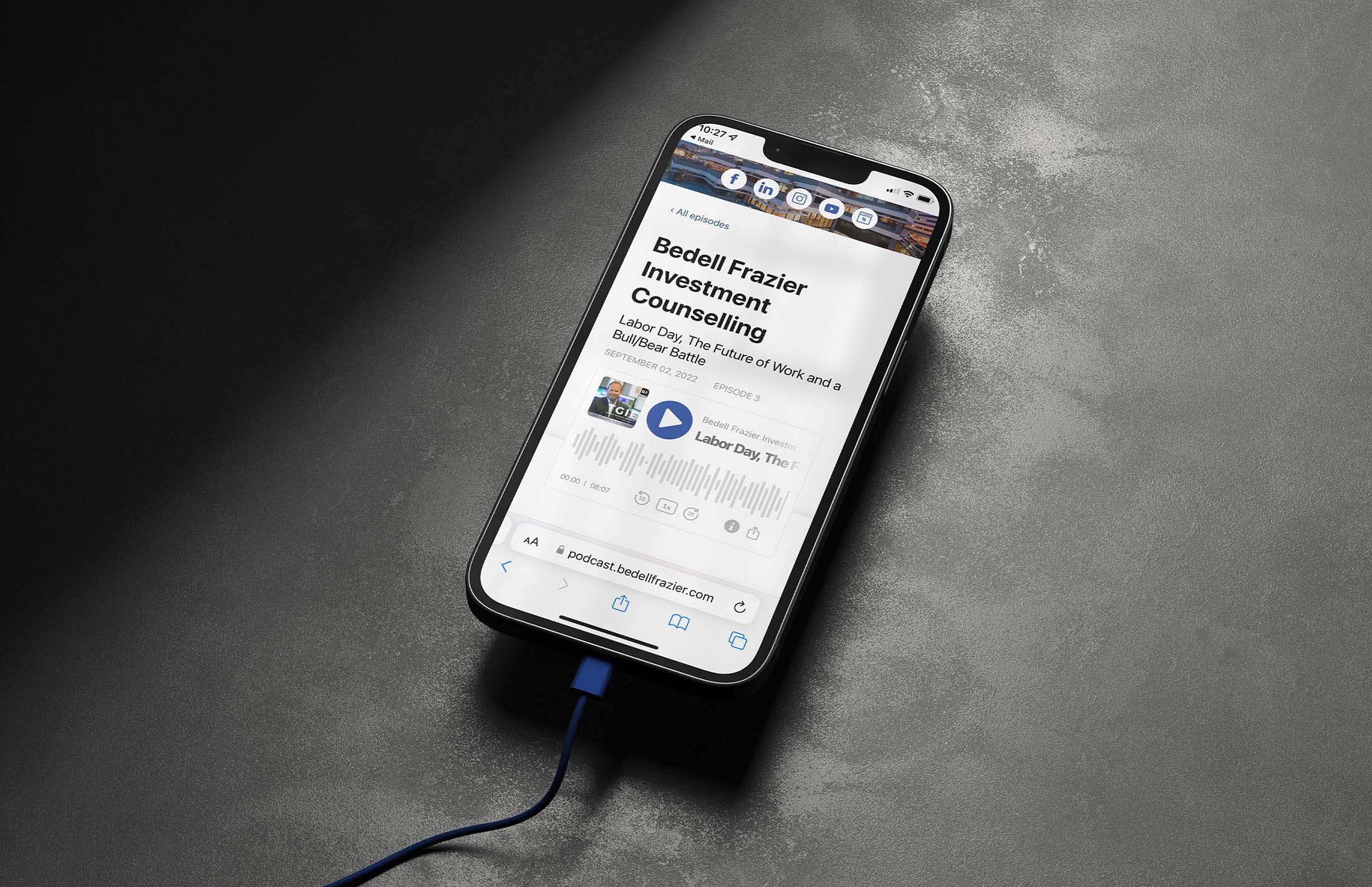 Smartphone displaying a Bedell Frazier Investment Counselling podcast episode titled ‘Labor Day, The Future of Work and a Bull/Bear Battle,’ connected via a blue charging cable on a dark surface.