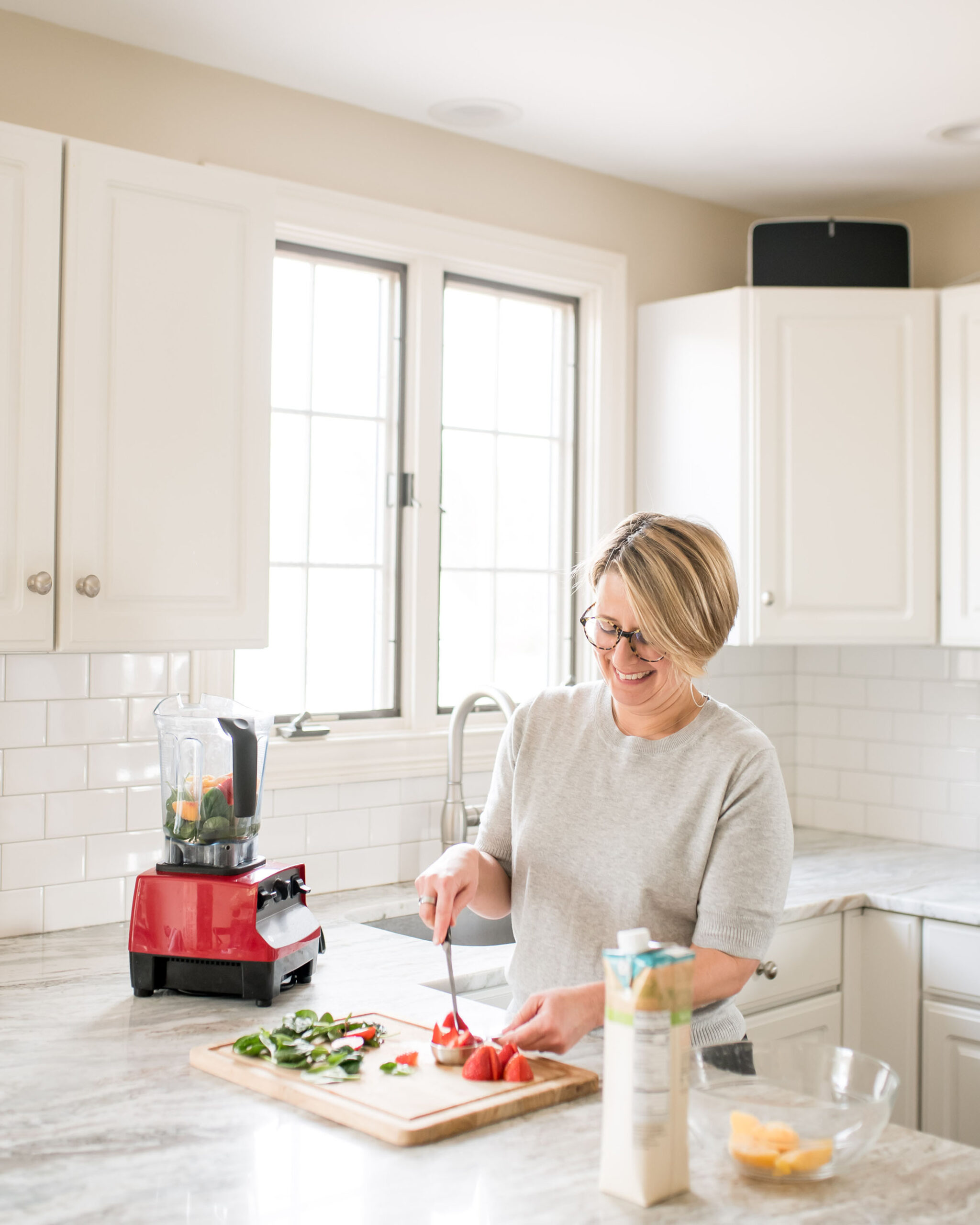 Jessica McManus in a bright kitchen, preparing fresh fruit and vegetables with a red blender on the counter, reflecting healthy living.