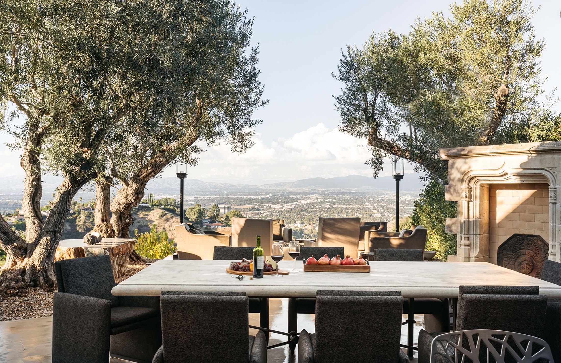 Elevated outdoor dining area with a modern table, chairs, and fireplace, overlooking a scenic city view framed by olive trees.