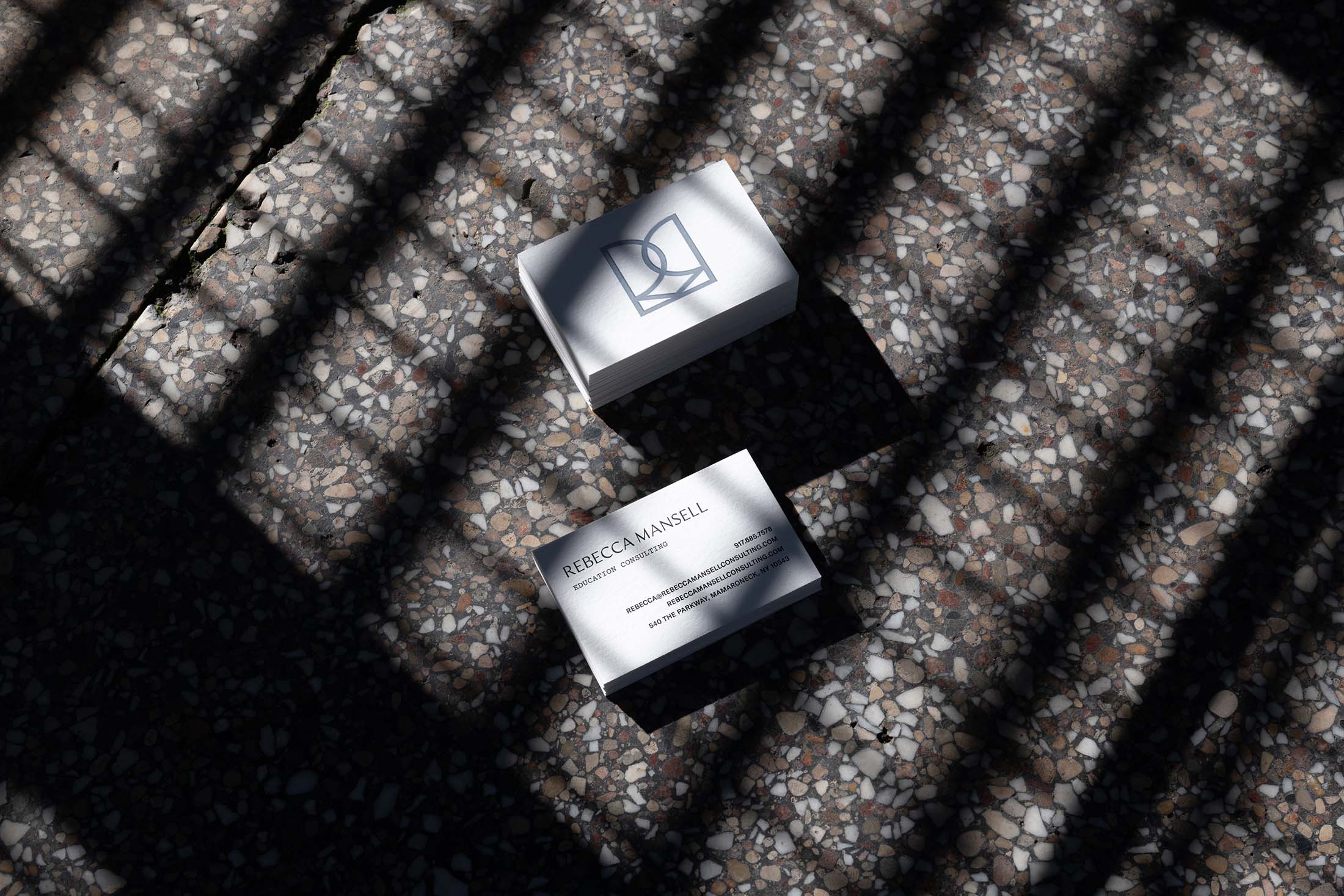 Rebecca Mansell business cards placed on a sunlit, terrazzo surface with shadow patterns, showcasing the brand’s clean, professional typography and logo.