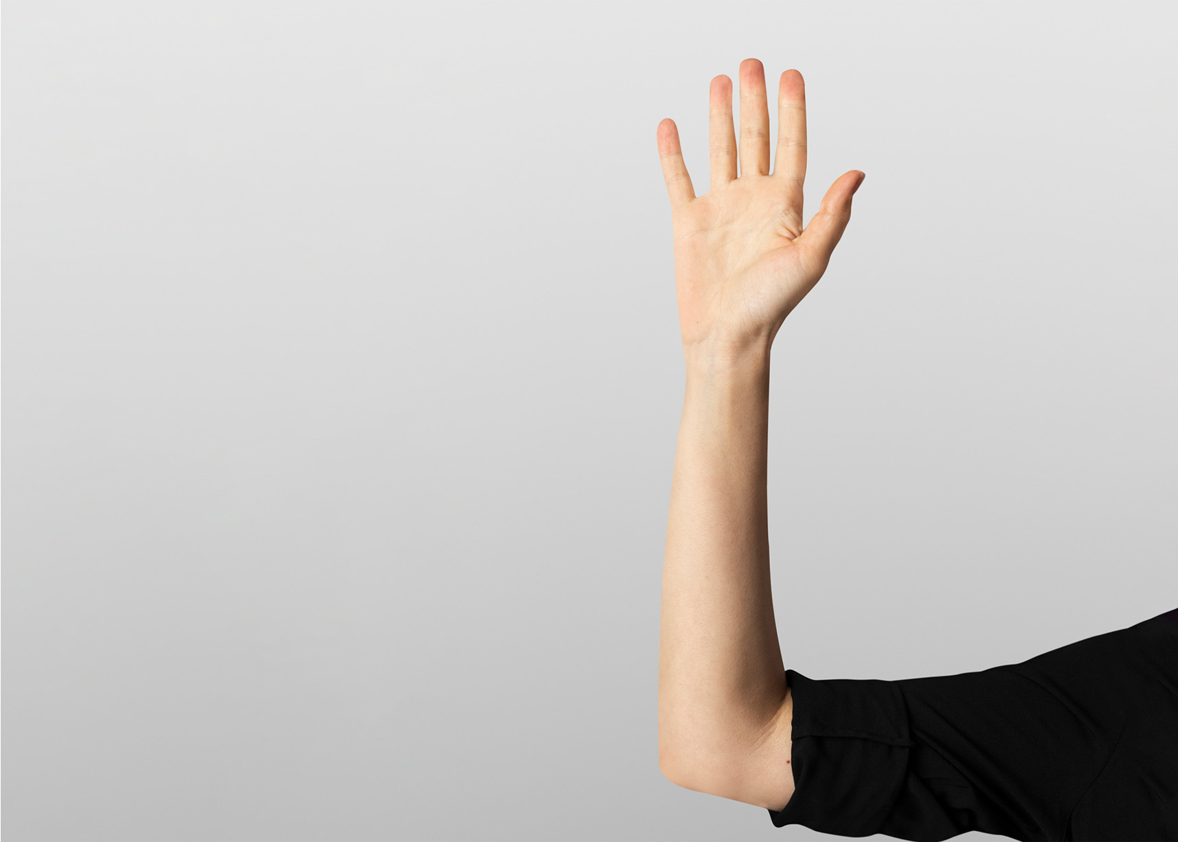 Raised hand of a person wearing a black shirt against a plain gray background, symbolizing volunteering or asking a question.