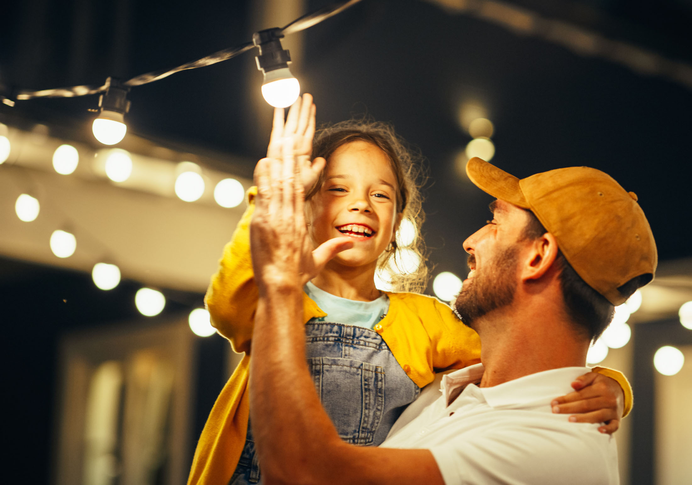 A smiling father holding his young daughter under string lights, celebrating together with a high-five in a warmly lit outdoor setting.