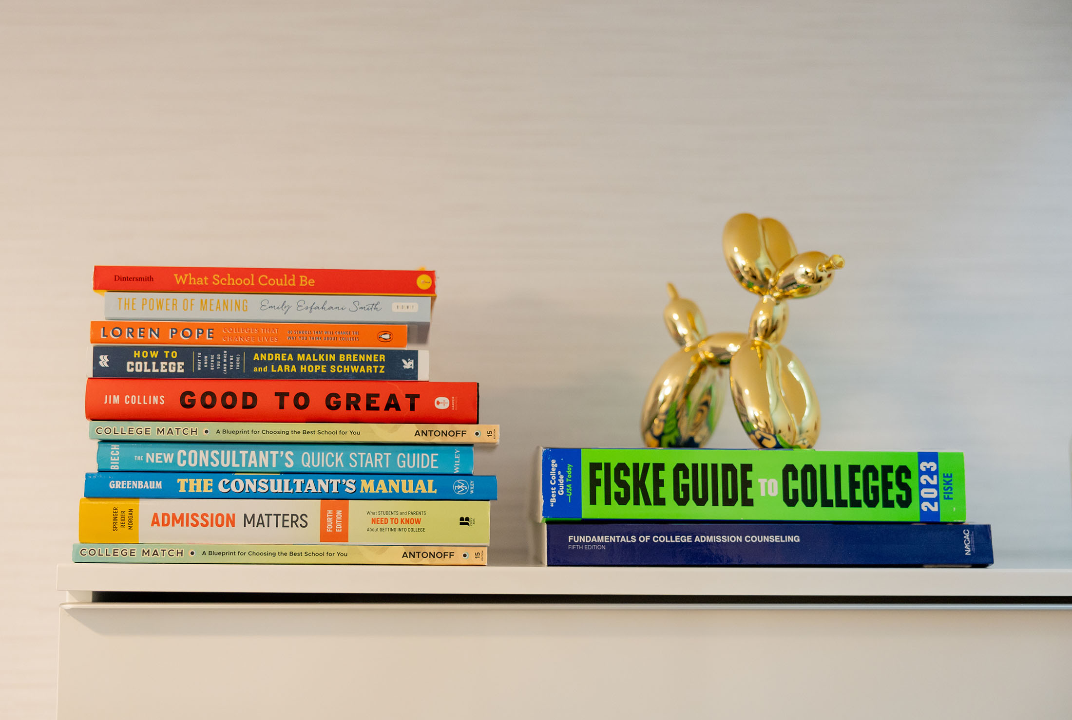 A curated stack of educational and consulting books, including titles such as “What School Could Be,” “Good to Great,” “The Consultant’s Manual,” and “Fiske Guide to Colleges,” accompanied by a golden balloon animal sculpture on a white shelf.