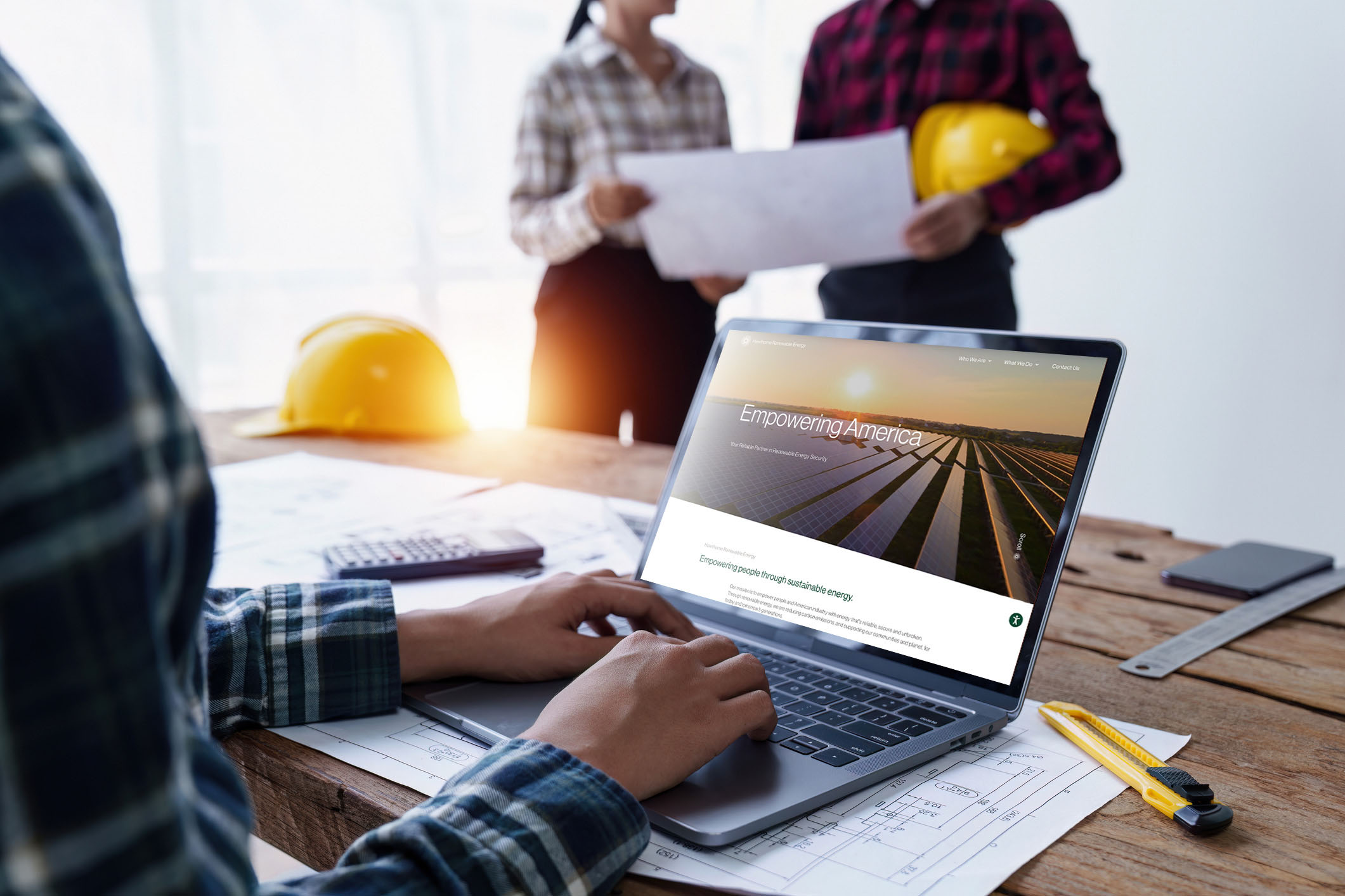 Close-up of a person using a laptop displaying the Hawthorne Renewable Energy website, with a worksite and two team members in the background.