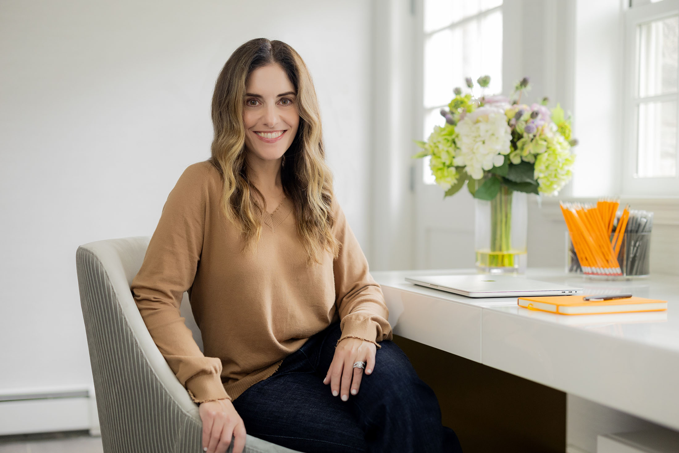 A welcoming portrait of Rebecca Mansell sitting in her bright and professional office, with a vase of fresh flowers and neatly arranged desk accessories in the background.