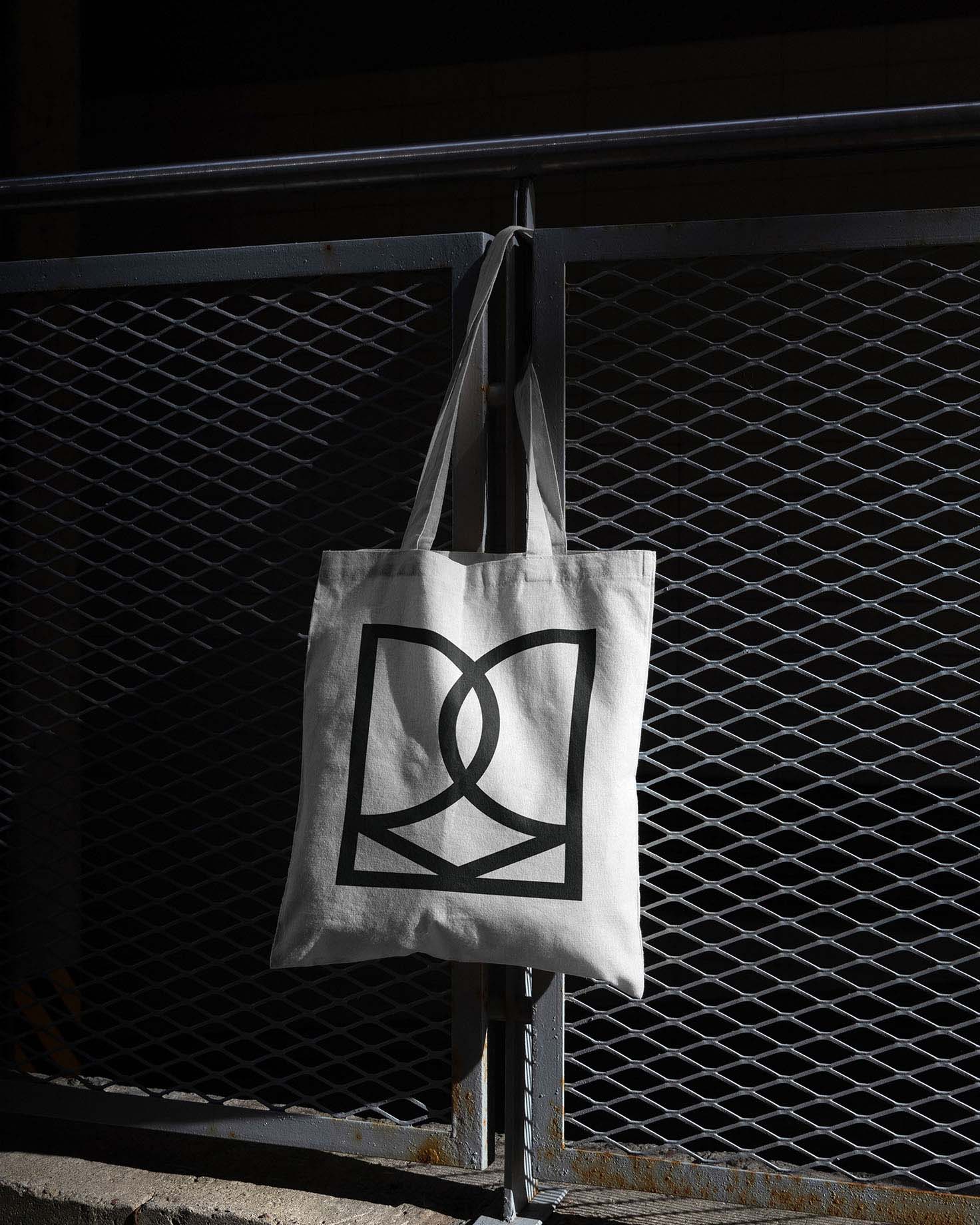 A white canvas tote bag with the Rebecca Mansell Consulting logo printed in black, hanging on a metal gate, captured with dramatic lighting.