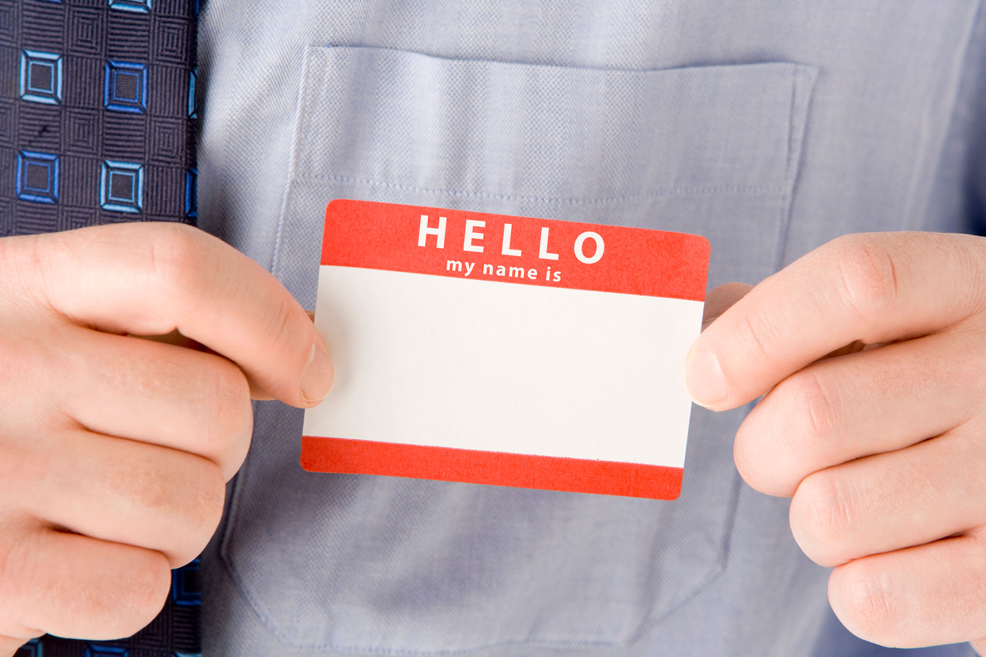 Close-up of a person in a light blue shirt holding a red and white name tag with ‘Hello my name is’ printed on it, leaving the name space blank.