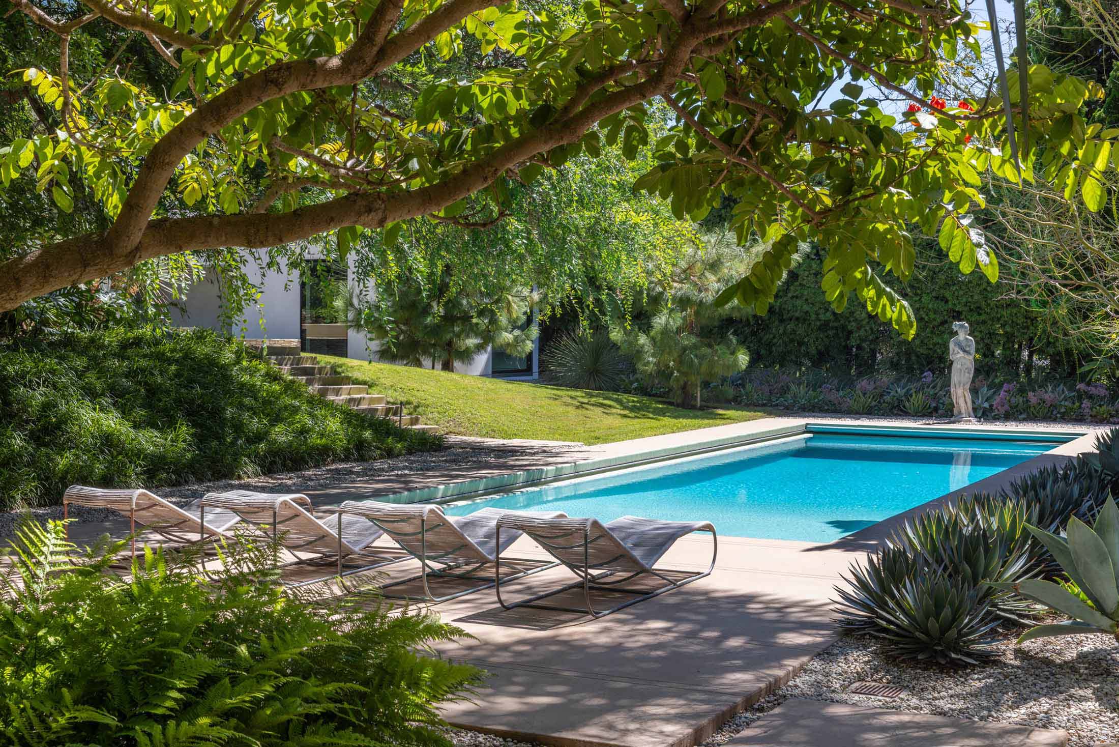 A serene backyard featuring a sparkling pool surrounded by lush greenery, modern lounge chairs, and a classic statue in a sophisticated landscape design.