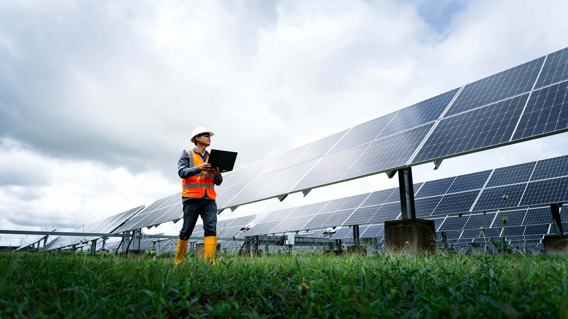 An engineer wearing a white hard hat, orange safety vest, and holding a laptop, inspecting a large solar panel field under a cloudy sky.