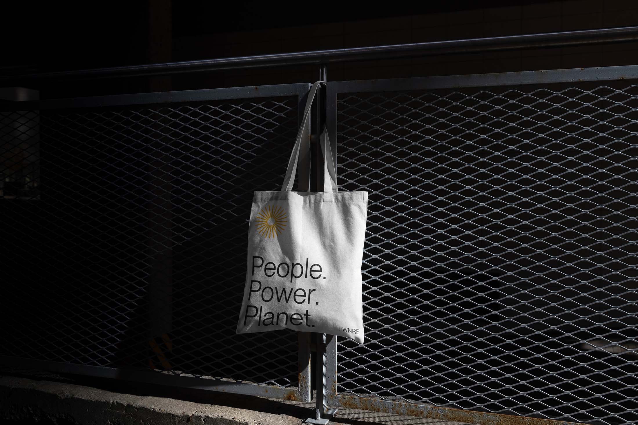 Hawthorne Renewable Energy branded white tote bag with the yellow sunburst logo and the text “People. Power. Planet.” hanging on a dark metal fence.