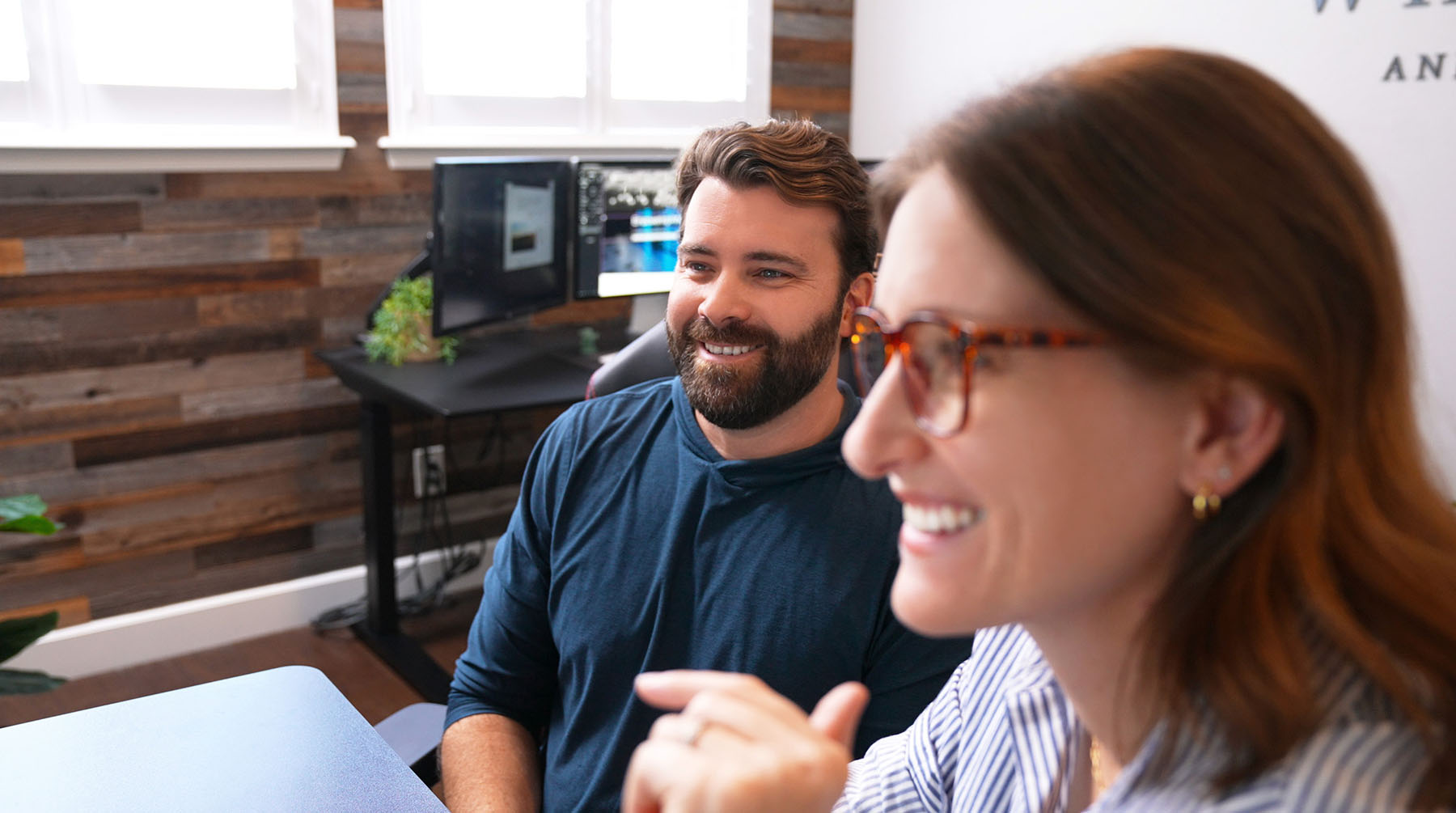 Steve Harris and Julie Harris of Whiskey & Red Smiling and Engaging in A Collaborative Discussion. the Modern Office Space Highlights Their Branding and Web Design Expertise, with Dual Monitors and A Wood-Paneled Accent Wall in The Background.