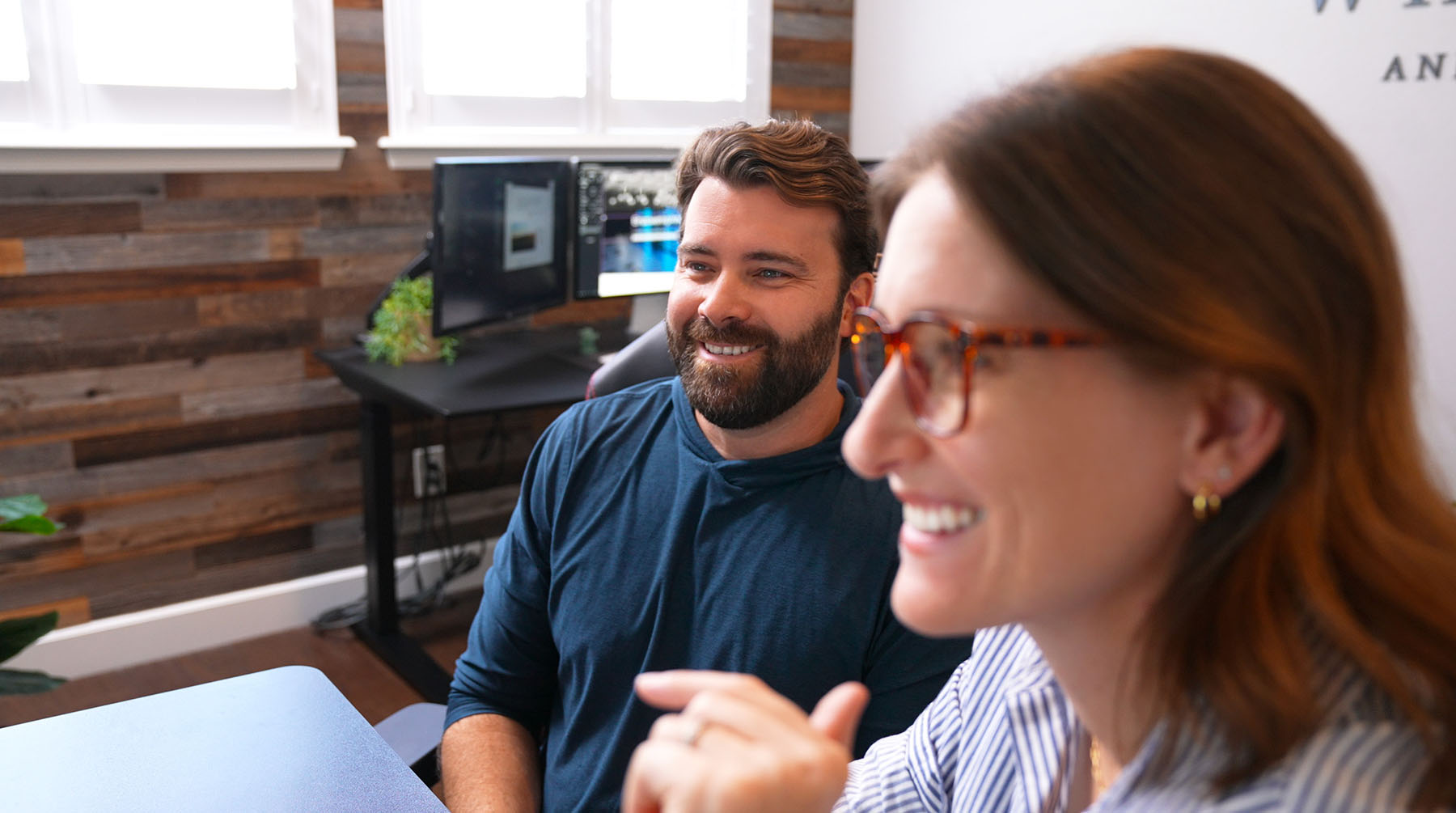 Steve Harris and Julie Harris of Whiskey & Red Smiling and Engaging in A Collaborative Discussion. the Modern Office Space Highlights Their Branding and Web Design Expertise, with Dual Monitors and A Wood-Paneled Accent Wall in The Background.