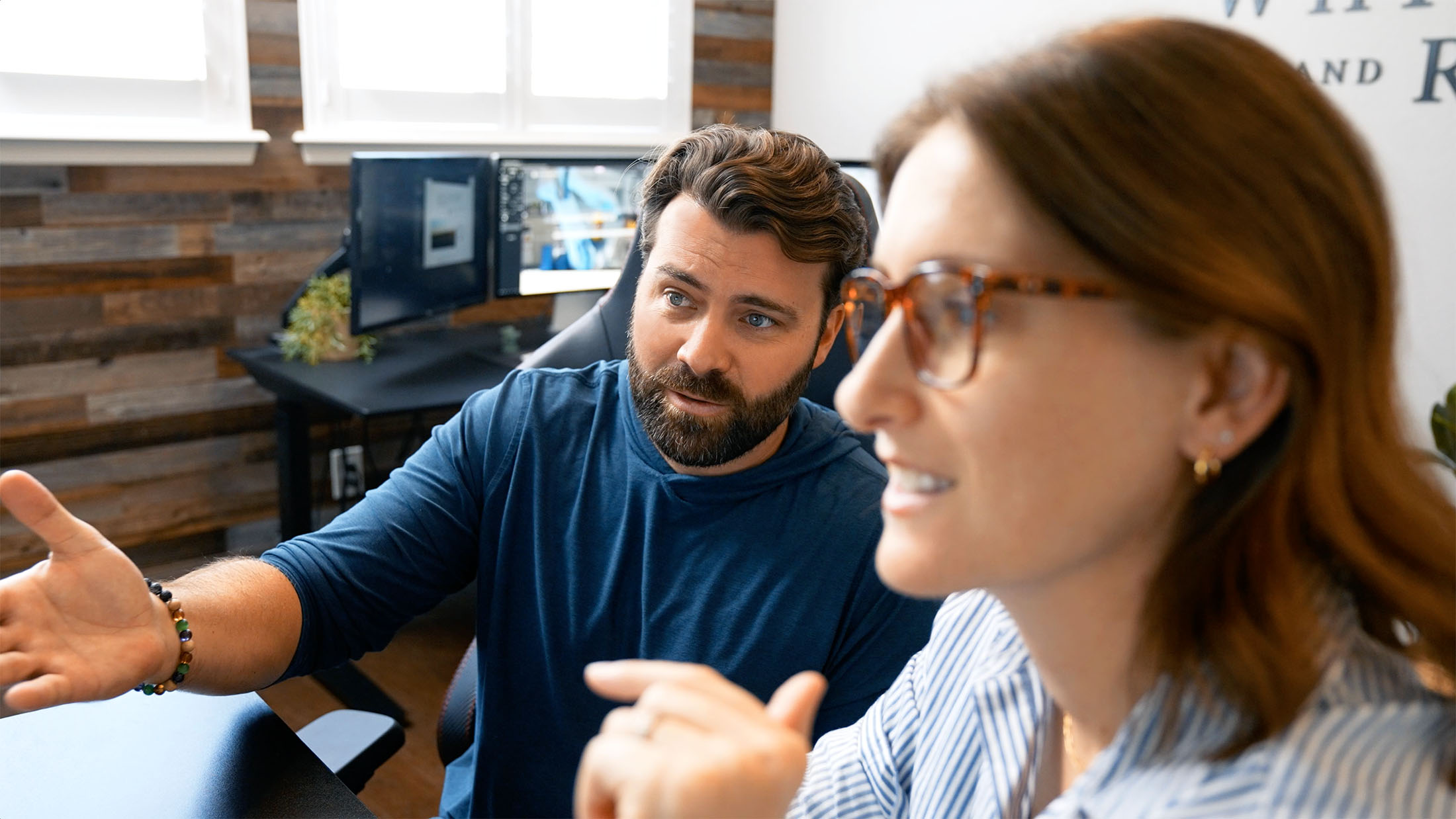 Steve Harris and Julie Harris of Whiskey & Red collaborating in a client-focused meeting, discussing branding and web design services, with a modern office backdrop featuring dual monitors and a wood-paneled wall.