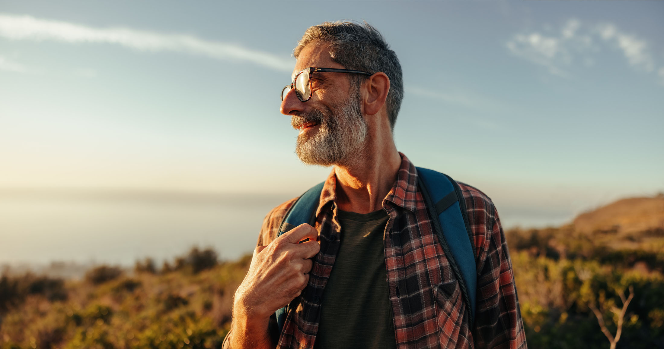 Close-up of a smiling man wearing glasses and a plaid shirt with a backpack, standing outdoors during golden hour with scenic hills in the background.