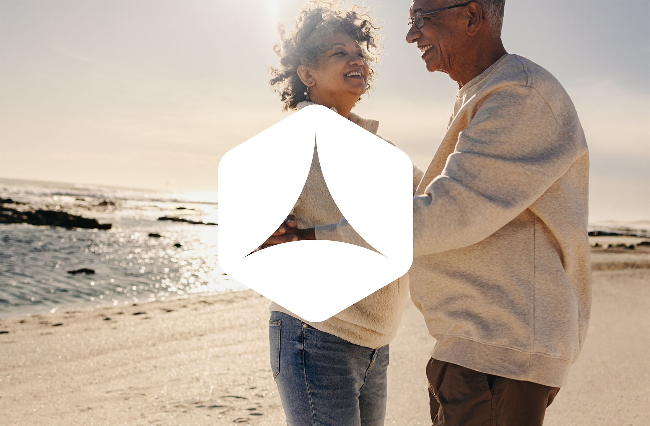 Smiling couple standing on a sunny beach, embracing each other, with the Stoic Private Wealth geometric logo overlayed in white.