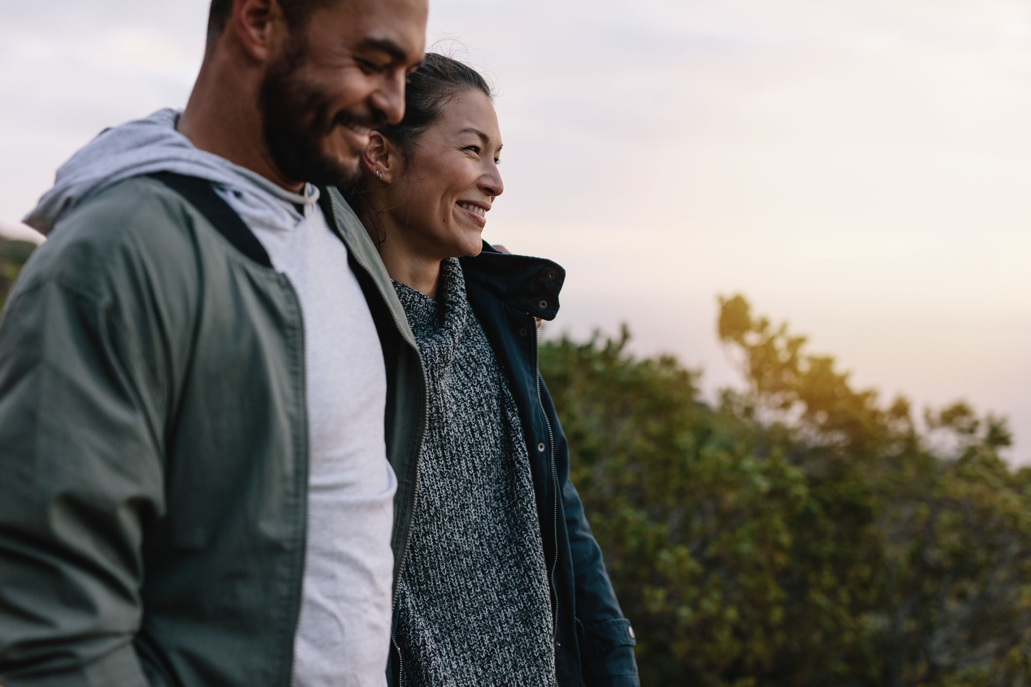 Smiling couple walking outdoors during sunset, dressed in casual, warm clothing with lush greenery in the background.