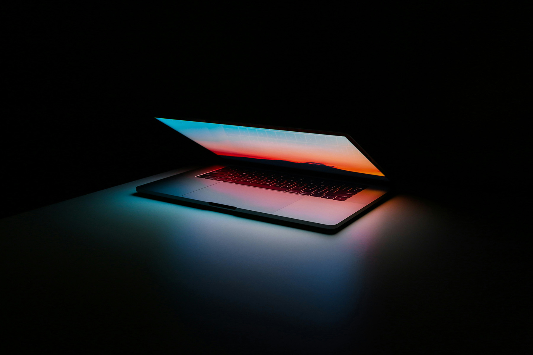 A sleek laptop partially open on a dark surface, illuminated by vibrant gradient lighting from the screen displaying warm sunset hues blending into cool tones, symbolizing modern technology and innovation.