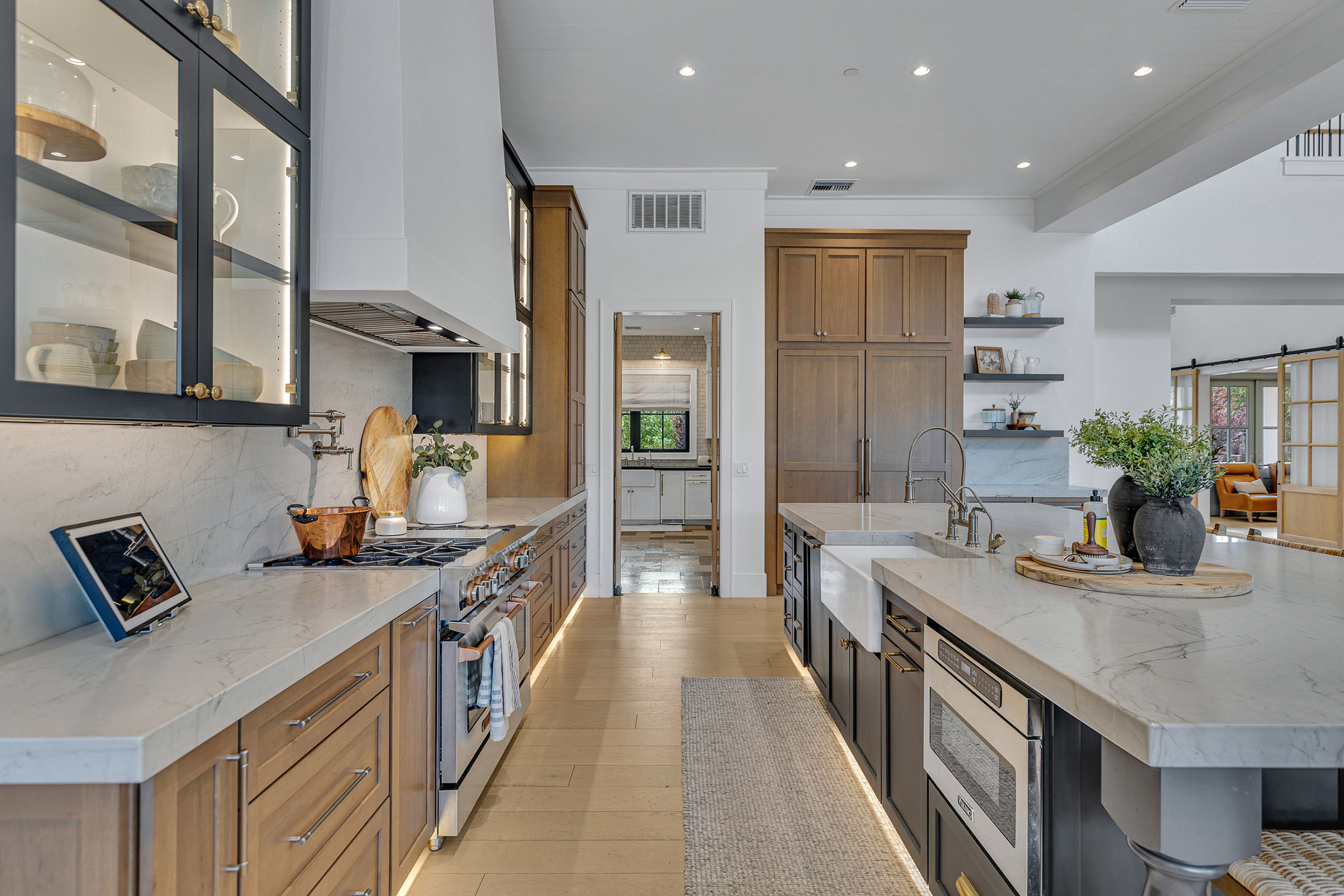 Spacious luxury kitchen featuring a blend of natural wood and dark cabinetry, marble countertops, a large island with farmhouse sink, high-end appliances, and warm ambient lighting, seamlessly flowing into an open-concept living space.