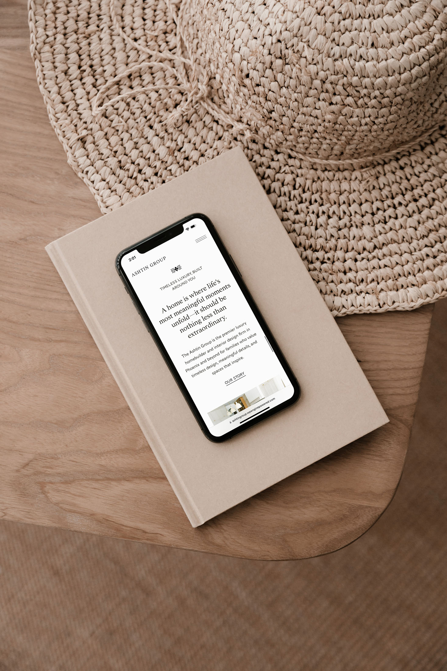 Smartphone displaying the Ashtin Group website homepage, placed on a beige hardcover book atop a wooden table, next to a woven straw hat, creating a warm and elegant aesthetic.