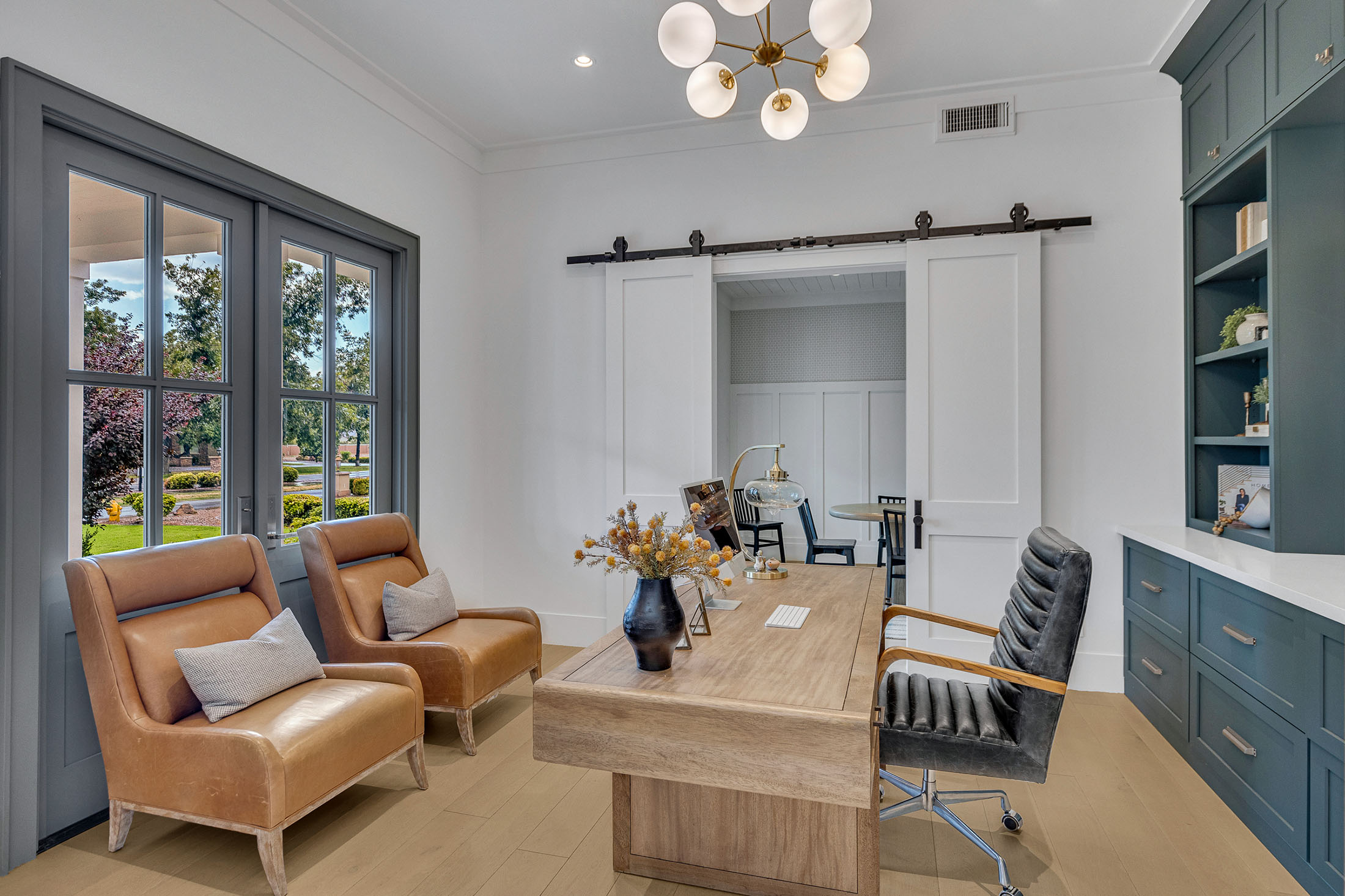 Elegant home office with a natural wood desk, leather chairs, dark green built-in shelving, and a modern chandelier, featuring large windows and a sliding barn door.