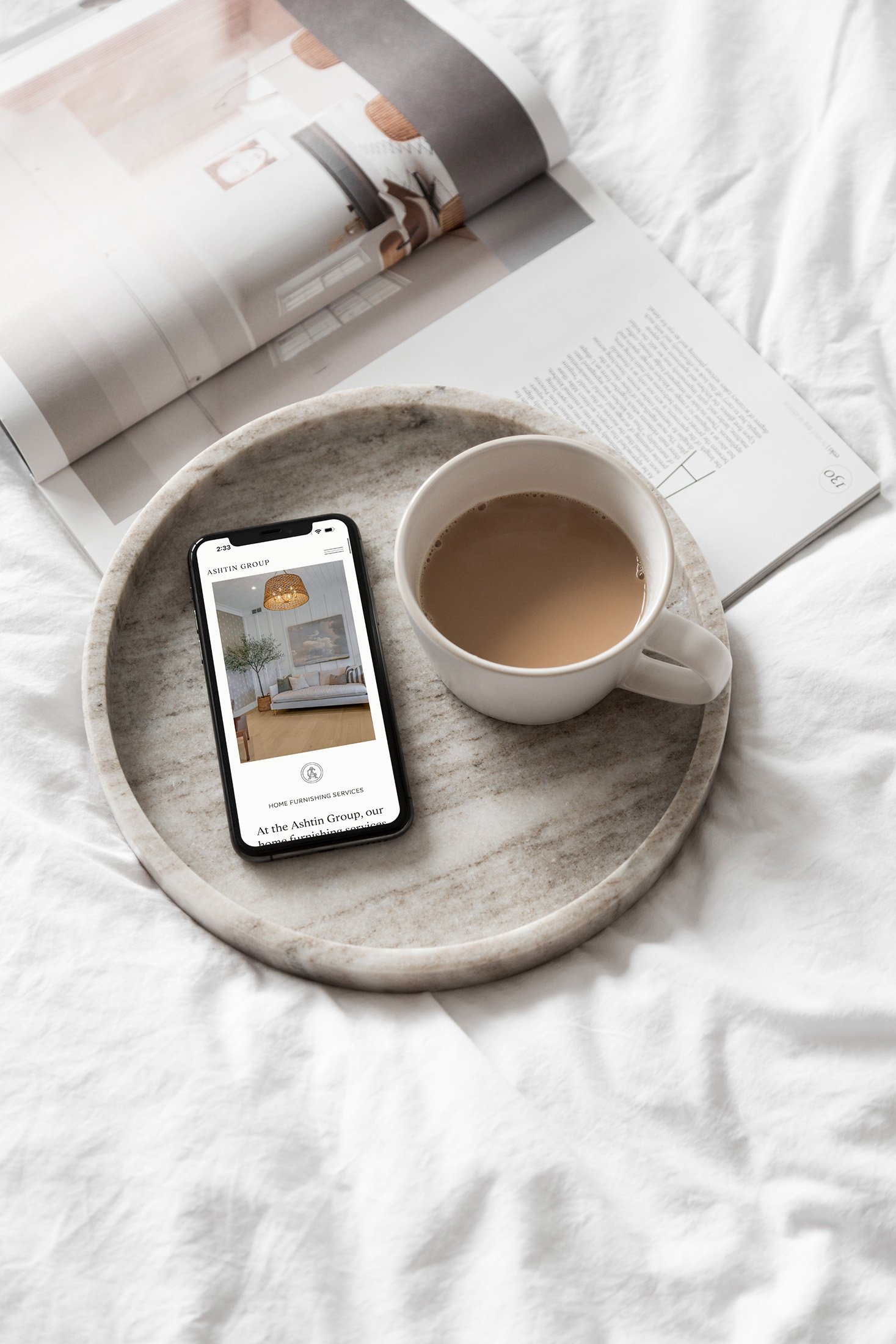 Smartphone displaying the Ashtin Group website on a marble tray with a cup of coffee, set on a white bed with an open interior design magazine.