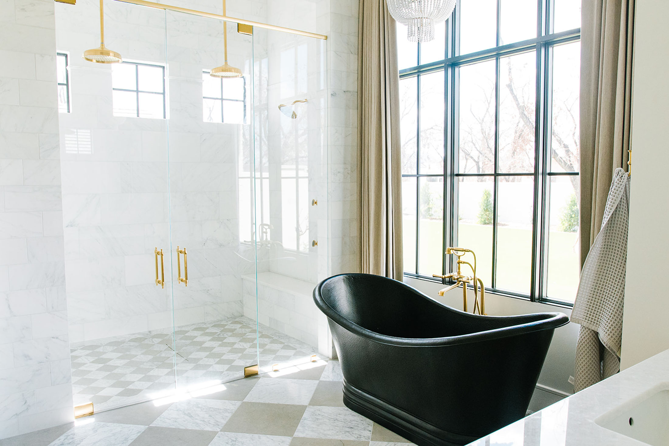 Luxurious bathroom with a black freestanding tub, gold fixtures, a spacious glass shower, and floor-to-ceiling windows with natural light.