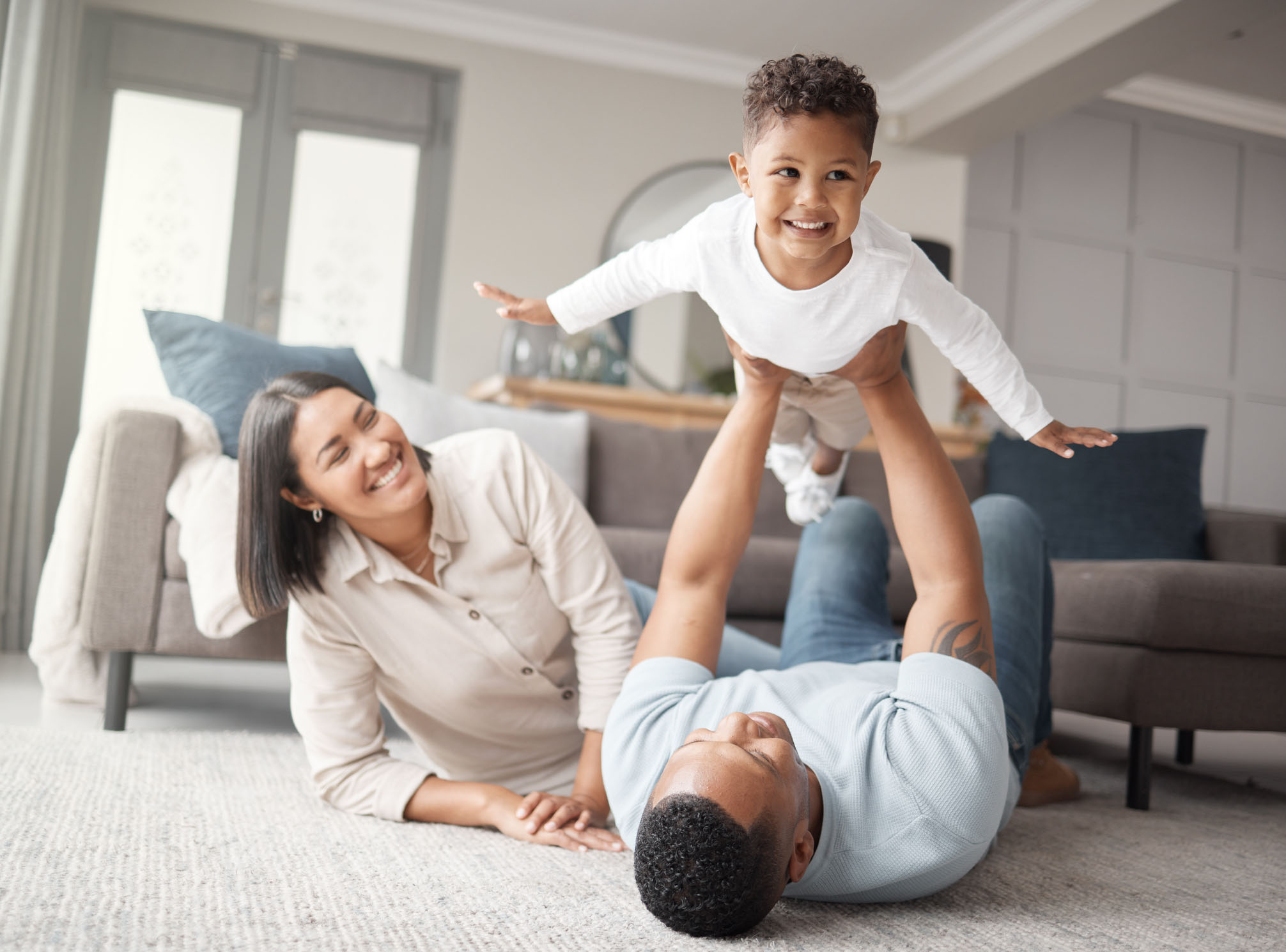 Smiling family playing together on a clean carpet in a bright living room, with a father lifting his child in the air and a mother watching joyfully, reflecting a safe and healthy home environment.