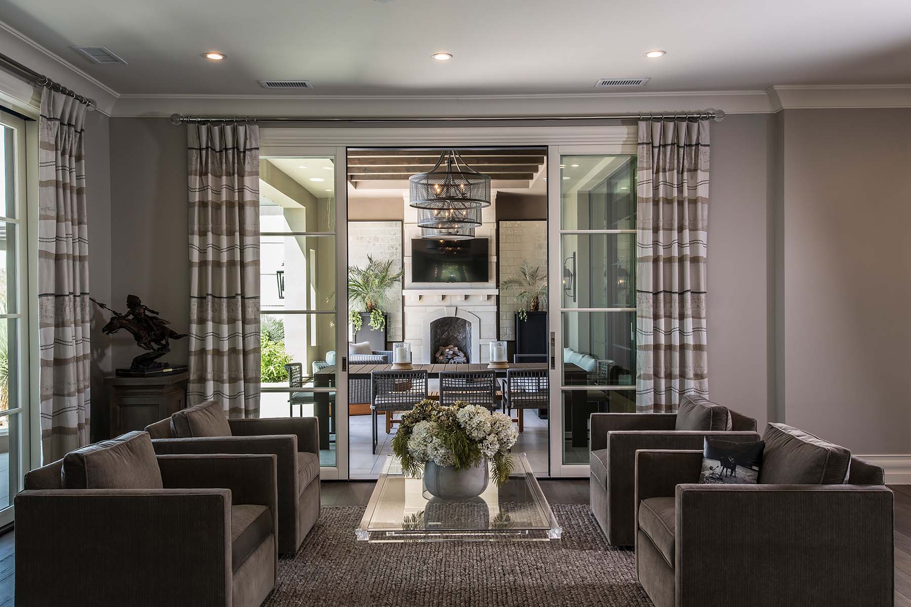 Elegant neutral-toned living room with velvet armchairs, a glass coffee table, and a view into a covered outdoor seating area with fireplace.