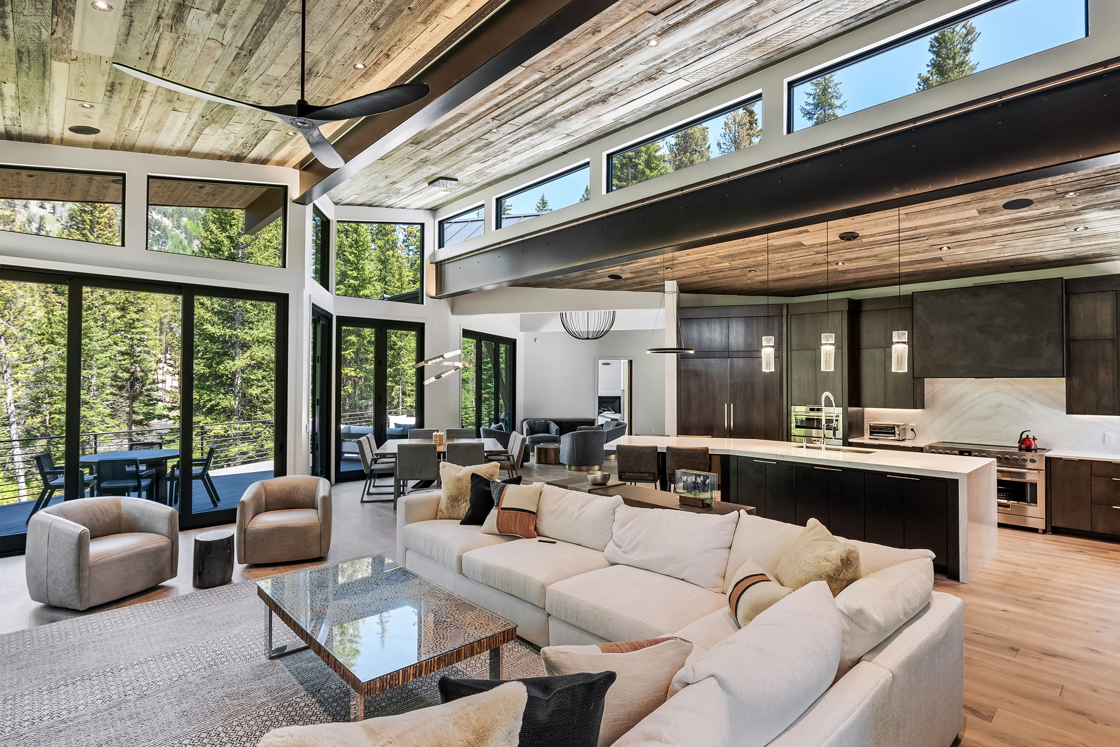Open-concept mountain home interior with modern kitchen, wood beam ceilings, large windows, and forest views.